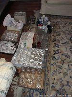 Full lot photo showing multiple trays and boxes filled with clear glass and crystal goblets, tumblers, pitchers, and assorted mugs arranged on a wooden floor next to a couch.