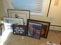 Five framed artworks arranged on floor leaning against wall and window, showing variety of sizes and frame styles.