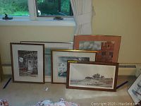 Five framed artwork pieces arranged on floor by a window, showing size and framing styles.