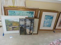 Seven pieces of framed artwork leaning against a wall, showing an assortment of landscape and water scenes in various frame styles and sizes.