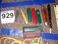 Lot of 7 pocket tools laid out on carpet with blue tape and measuring tape, includes Swiss Army style knives and other folding knives, with sheaths included.