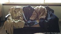 Four coats and jackets laid out on chairs and sofa including beige trench coat, black leather coat, faux fur and suede stole jacket, and black cashmere coat.