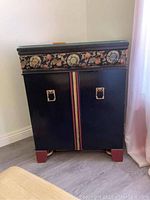 Front view of closed black vintage sewing cabinet with decorative floral and medallion trim and metal door handles.