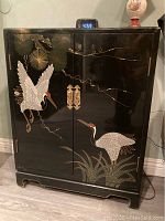 Front view of black lacquer cabinet with hand-painted crane and botanical motif, digital clock on top and lamp to the right.