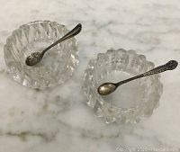 Two glass salt cellars with sterling silver spoons resting inside them on a marble surface.