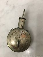 Front view of the oval metal perfume case showing the leaf engraved design and the attached stopper/spoon applicator.