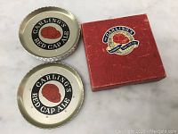 Two metal Carling's Red Cap Ale coasters with embossed edges and printed logo, alongside original red box with 1951 International Trade Fair label.