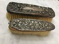 Two antique hair brushes with silver backs showing ornate floral and scrollwork designs and natural bristles on wooden bases.