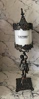 Full front view of the decorative vintage metal piece, highlighting the white cylinder with 'CAUVERY' label, metal floral designs, and figurine stand.