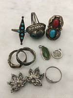 Clear image of the sterling silver jewelry pieces laid out on a light surface showing rings with turquoise and red stones, floral clips, green gemstone pendant, and a round charm.