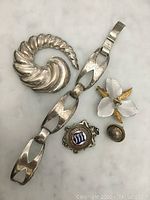 Top view of the five sterling silver jewelry items including large shell brooch, linked bracelet, floral brooch, round button and decorative enamel badge pin