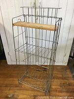 Full view of wire display shelf showing metal wire frame and wooden accent plank, placed on wooden floor against white paneled wall.