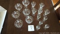Overhead view of 6 vintage crystal fruit nappies and 10 stemmed glasses arranged on a dark wood surface, showing etched floral patterns.