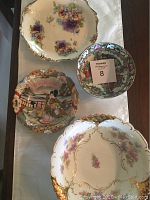 Four vintage oriental style decorative plates and bowls arranged on a white cloth surface, showing detailed floral and oriental painted designs with gold embossing and scalloped edges.