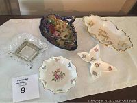Five assorted vintage decorative dishes on white tablecloth, including cobalt blue and gold leaf-shaped dishes, floral porcelain dishes, and clear glass dish.