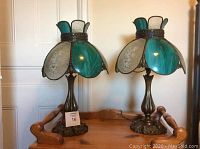 Photo of two Tiffany-style lamps with brass bases and emerald green and etched clear glass shades placed on a wooden surface.
