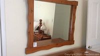 Front view of rectangular wall mirror with natural pine frame mounted on wall above dresser, showing reflection of room including bed and lamp.