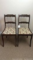 Front view of two identical wooden antique chairs with floral cushioned seats, showing carved top rails and wavy slats on the backrest.
