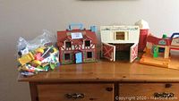 Three vintage Fisher Price plastic playsets: a Tudor style dollhouse, a white barn with red doors and silo, and an orange garage/playset with a blue slide handle. A clear plastic bag of small Fisher Price vehicles and figures is also shown.