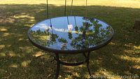 Round glass patio table with metal frame and legs, positioned outdoors on grass.