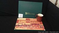 Photo of green Eatons Recollections box, one placemat laid on top showing vintage street scene with horse-drawn carriages, and one brown mug on couch