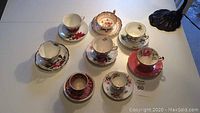 Photo of 6 assorted vintage tea cups and matching saucers arranged on a table showing various floral patterns and gold trim