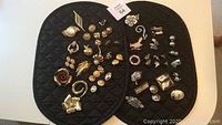 Two black quilted mats with an assortment of vintage brooches and clip earrings arranged on them. Items are in gold and silver tones with various designs.