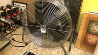Front view of the floor fan showing the metal grille, frame, and black blades with lot number tag attached.