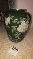 Front angled view of green glazed ceramic vase with white floral and leaf decoration and gold rim accents.