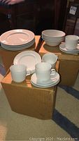 Overview of white dinnerware items arranged on three cardboard boxes, showing stacks of dinner plates, bowls, and cups on saucers