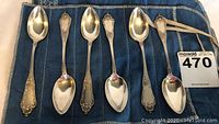 Top down view of all six silver tea spoons arranged side by side in a blue cloth holder. The spoons have ornate handles and a hallmark stamped on edges.