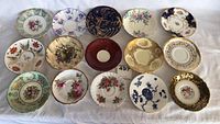 Full overhead view of all fifteen tea saucers laid out on white cloth, showing various floral and patterned designs in multiple colors and gold trimming.