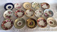 Full view of 15 assorted tea saucers arranged in rows on white background showing varied floral and gold designs.
