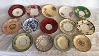 Overview photo showing all 14 tea saucers with various floral and gold designs, different colors and styles visible