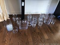 Photo showing assortment of 14 clear glass vases of varying heights and shapes arranged on hardwood floor, along with one container of clear decorative filler beads
