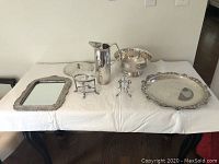 Full overview of all six items displayed on a white covered table including tray, mirror frame, candy dish stand, pitcher, bowl, and bud vase.