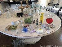 Overall view of various glass items and decorative pieces arranged on round marble table including Orrefors candle holder, glass vases, paperweights, BODA glass bowl, glass apples, candles, mirror, and picture frame.