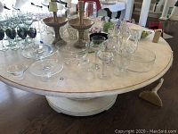 Overall view of the lot on a round table including various glassware items: three-tiered stand, glass dome, candle holders, pitcher, and Design Guild bowl.