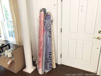 Seven rolls of various upholstery fabrics leaned against a wall near a white door and column. Some rolls have protective plastic calico sleeves.