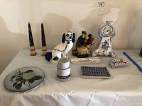 Overview of the entire lot showing all items arranged on a table covered in white fabric. Items include two candlesticks, ceramic dog, balloon seller figure, hand painted clock, various dishes and plates.