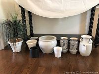 Full view of the lot showing seven ceramic vases of various sizes and designs arranged on a wooden floor under a table.