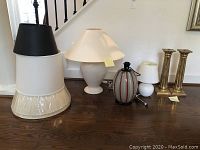 Group photo of lamps, lamp bases, and lamp shades arranged on floor including white French lamp, striped glass lamp, round white lamp, multiple shades.