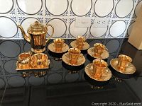 Full view of the complete gold tea set on black surface: teapot, sugar bowl, creamer, six teacups and saucers arranged.