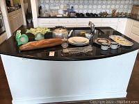 Full view of all items included in the lot placed on a black kitchen island including plates, teapot, cups, tray, pedestal dish, wooden bread, and picture frame note holder.