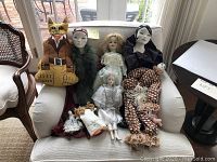 Wide view of two handmade dolls including a large stuffed cat, multiple porcelain and cloth dolls arranged on a white chair.