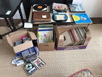 Overview of three cardboard boxes containing vinyl records, CDs and DVDs on floor