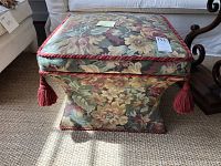 Front view of the tapestry fabric ottoman showing floral pattern upholstery and red tassels on corners.