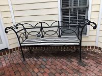 Front view of black iron outdoor bench with black and white striped cushion on brick patio.