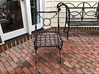 Front and side view of black iron chair with lattice iron seat and decorative backrest, set on brick flooring outdoors