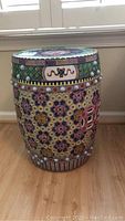 Full view of cylindrical ceramic stool showing multicolor floral and geometric pattern, raised knobs and Greek key border.
